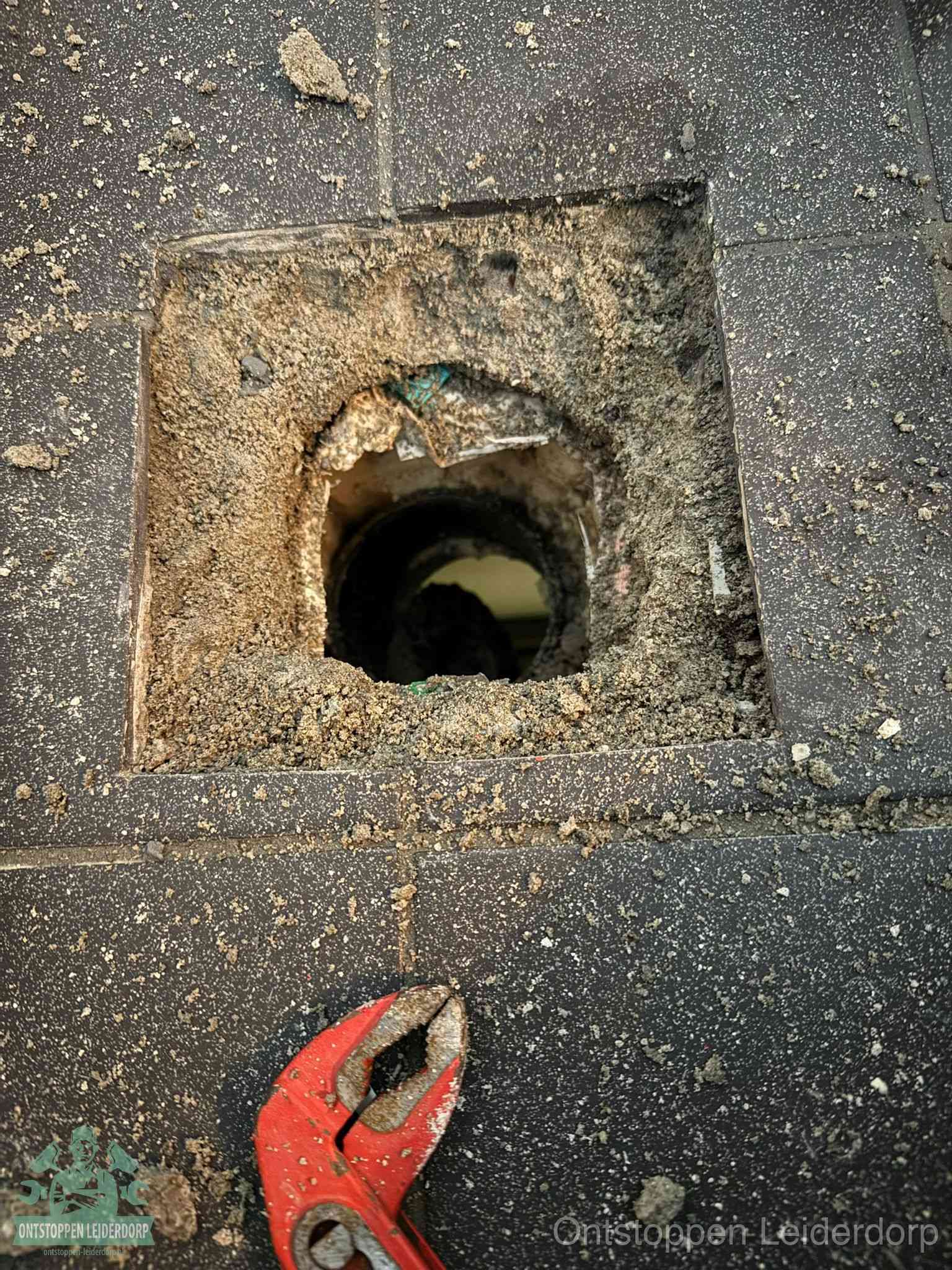 Een geopend rioolputje in een betontegelvloer met losgemaakt rooster en een zichtbare ronde afvoerbuis, het achterhalen waar de afvoerverstopping zit in Leiderdorp