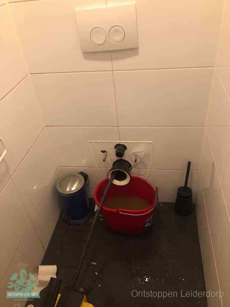 Een ontstopte wand-wc met verwijderde toiletpot waar een ontstoppingsveer in de wanduitgang verdwijnt, het achterhalen waar de verstopping zit in Leiderdorp