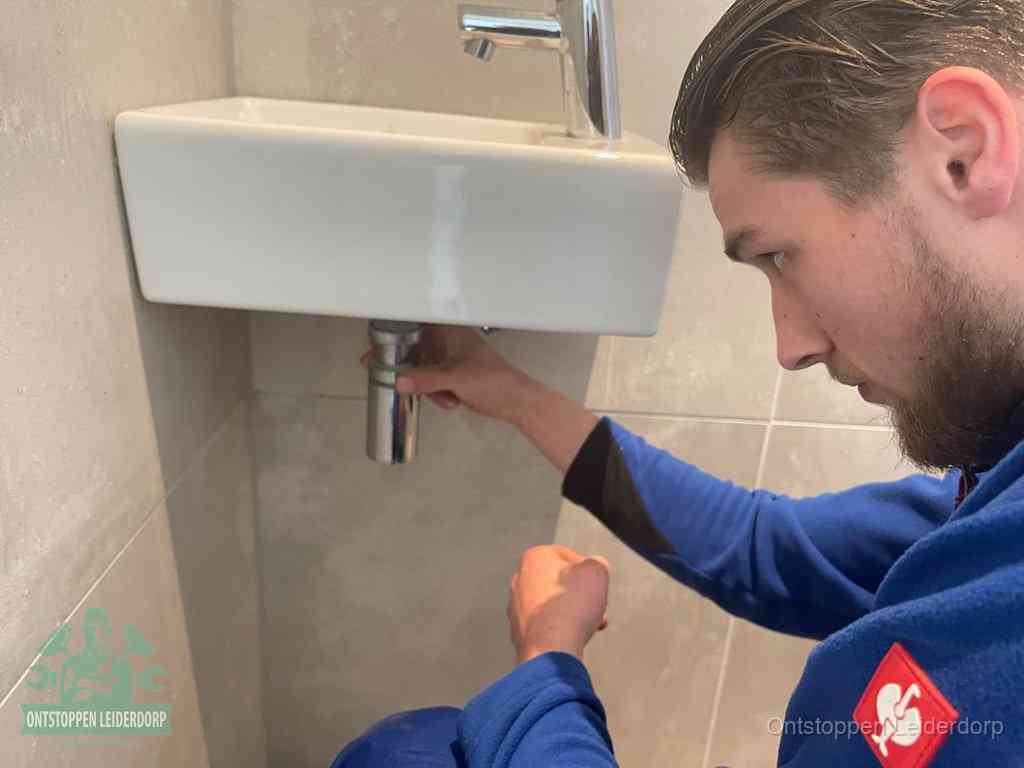 Een monteur draait met de hand de zwanenhalssifon onder een fonteinwasbak in de toiletruimte los om de bron van rioollucht in Leiderdorp te vinden