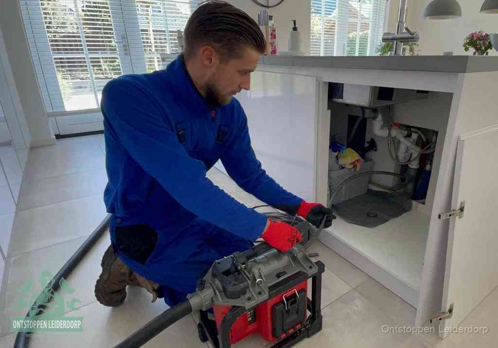 Een monteur voert een professionele elektrische ontstoppingsmachine in de afvoer onder de spoelbak tijdens het gootsteen ontstoppen in Leiderdorp